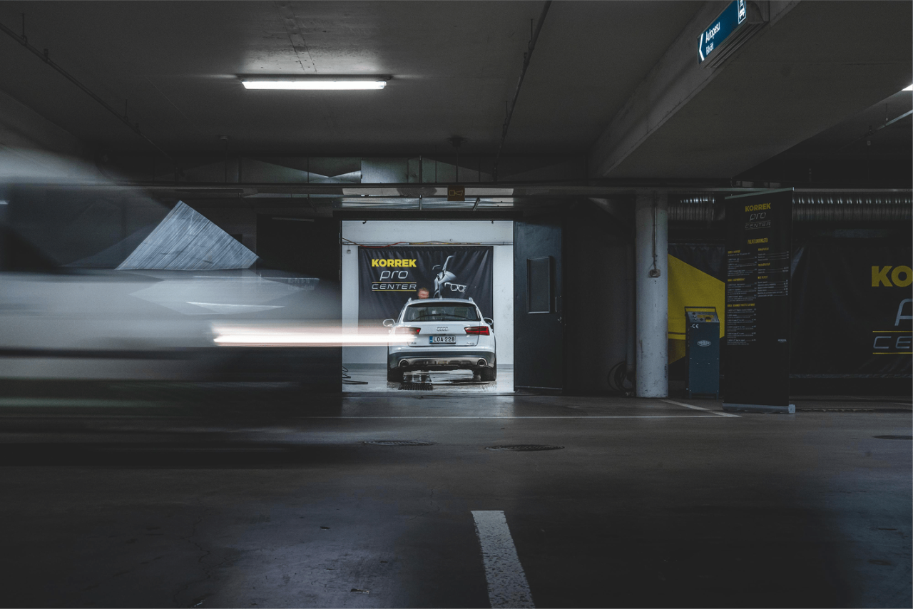 Pysäköintihallissa näkyy KORREK Pro Center -pesutila, valkoinen auto sisällä ja ohi ajavan auton liike-epäterävyys etualalla.